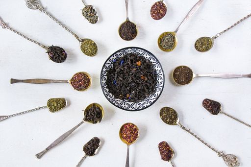 Flat lay of an assortment of dried tea leaves in a ceramic saucer and vintage silverware spoons on a white background