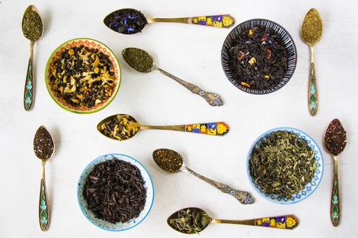 A top view of herbal and natural dry tea set in colorful small bowls and vintage silverware spoons on a white background