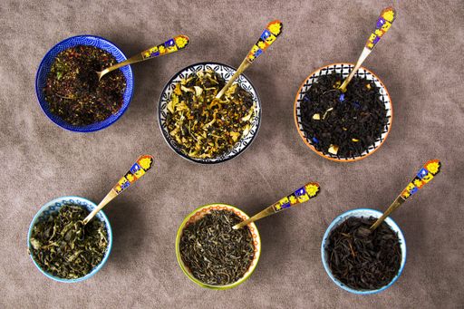 A top view of an assortment of herbal and natural dry tea in colorful small bowls and vintage silverware spoons