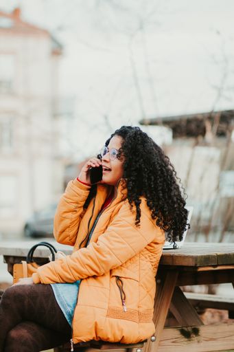 A vertical shot of a young curly woman sitting on a bench while talking on her mobile phone in the park