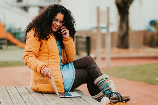 A young curly woman sitting on a bench using laptop while talking on a phone in the park
