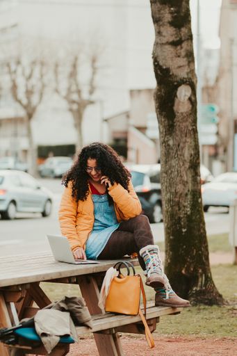 A vertical shot of young curly woman sitting on a bench using laptop while talking on a phone in the park