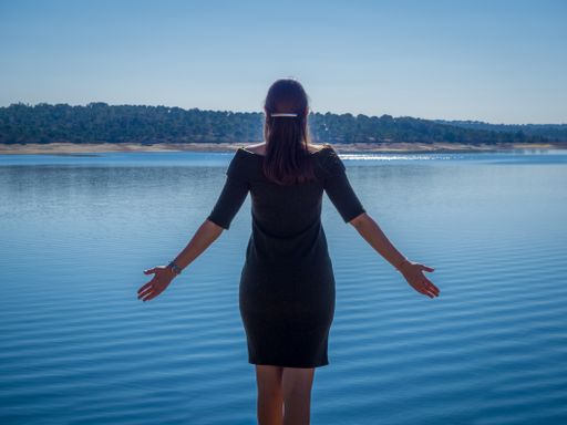 A young female in a nice black dress standing with her arms open enjoying the beautiful morning view of the sea