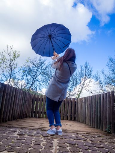 A vertical shot of a female holding a blue umbrella on a bridge surrounded by leafless trees