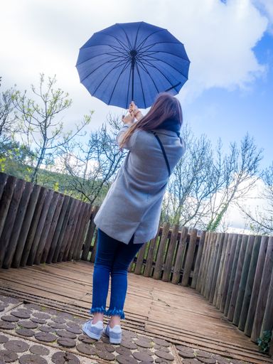 A vertical shot of a female holding a blue umbrella on a bridge surrounded by leafless trees