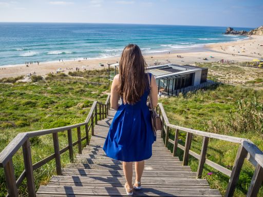 A young long-haired female in a beautiful blue dress walking down the stairs heading to the beach