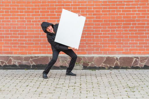 A man in black clothes stands against a red brick wall and holds a blank white canvas in front of him to paint