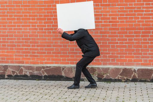 A man carries on his shoulder a blank white canvas for painting against a red brick wall