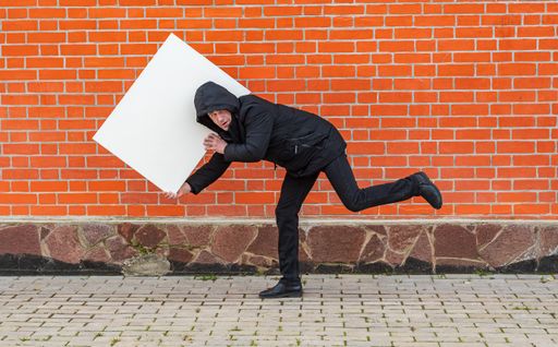 A man in black clothes is bent under the weight of a painting canvas against a red brick wall and looks into the camera