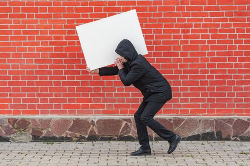 A man carries on his shoulder a blank white canvas for painting against a red brick wall