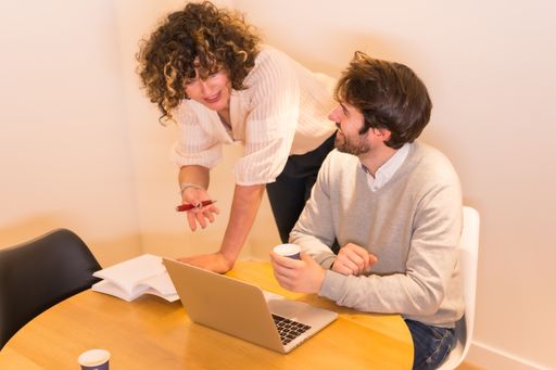 A female boss giving directions to the worker at a table in the office, with a computer and a notepad