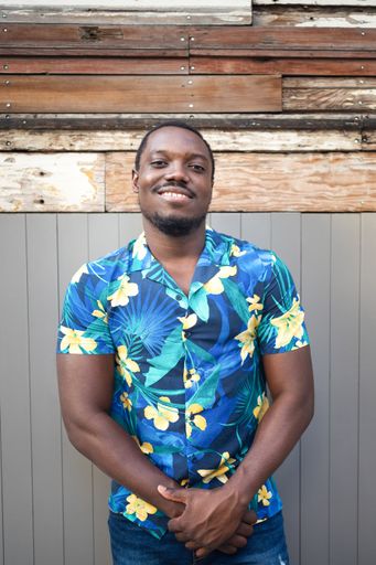 A vertical shot of a handsome black man in a blue floral shit, posing happily outdoors