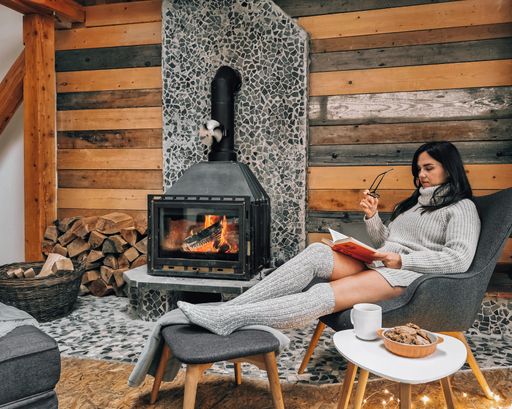A woman is sitting with a cup of hot drink and a book near the fireplace
