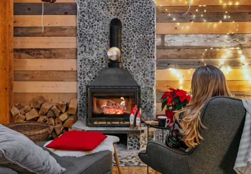A woman is sitting with a cup of hot drink and a book near the fireplace