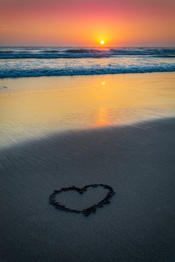 A vertical shot of a beautiful sunset at the beach and a drawing of a heart on the sand