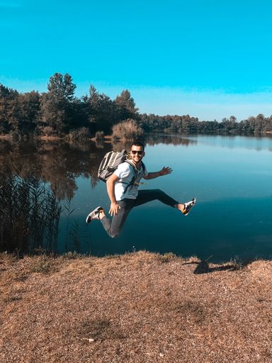 A shallow focus of an excited young male with a backpack jumping on the shore of a lake