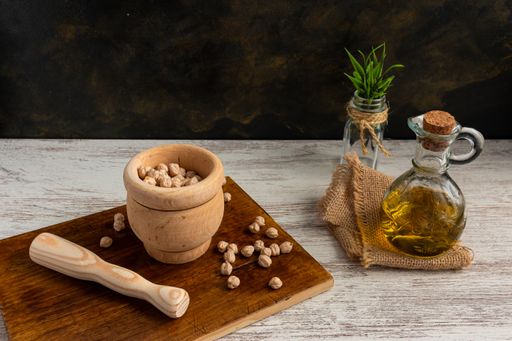 A closeup of chickpeas in a wooden mortar and pestle, next to an olive oil bottle, on a wooden surface