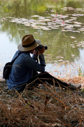 A man with the camera sitting on the grass by the water.