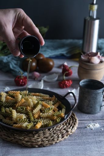 A cook's hand pouring oil in the fusilli pasta pot with fresh ingredients for cooking on a wooden rustic table