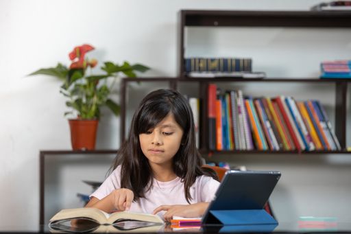 A Hispanic schoolgirl reading a book and using a tablet at home - online education
