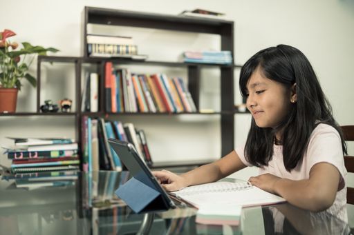 A happy Hispanic schoolgirl doing a homework using a notebook and tablet at home - online education