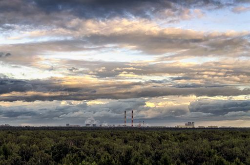 A beautiful overhead shot of a thick forest and a breathtaking cloudy sky with factory pipes visible in the distance