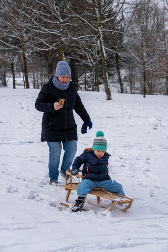 Woman taking a photo with her smartphone of boy sitting on wooden sled on a snowy hill at the park on a cold winter day.