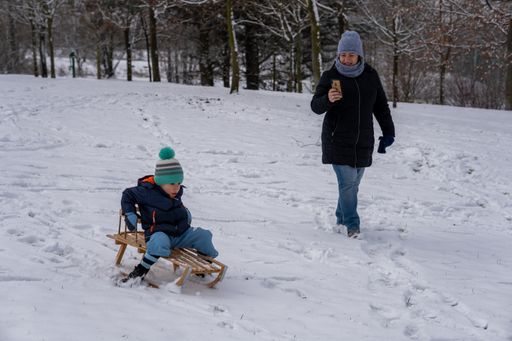 Woman taking a photo with her smartphone of boy sitting on wooden sled on a snowy hill at the park on a cold winter day.