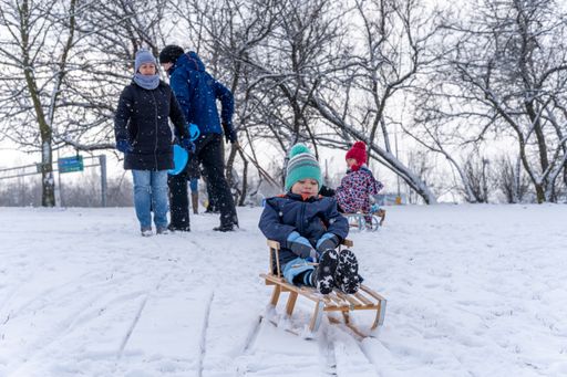 Young boy sitting on a wooden sled on snow hill while adults are watching at a park on a cold winter day.