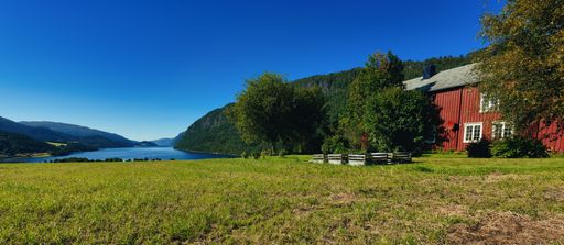 Panoramic view at a norwegian fjord with a small red farmhouse against a cloudless clear blue sky and a green meadow and some trees in the garden