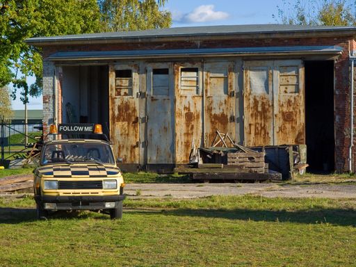 A vintage rusty yellow pickup truck is parked in front of a rustic wooden barn in a rural setting