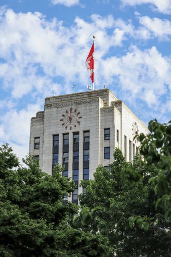 A vertical shot of the Vancouver City Hall with the Canadian flag seen behind lush green trees