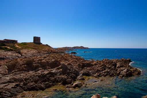 A wide shot of a rocky hill with a castle building near the open sea in a clear sunny blue sky in the background