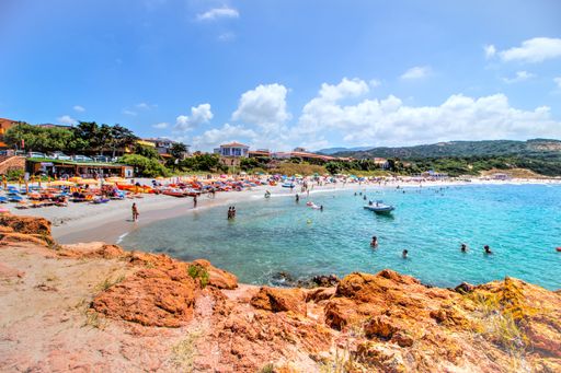 A wide shot of a sandy beach resort full of people, surrounded with mountains in a clear sunny blue sky