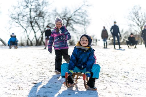 Young boy on sled and running girl with parents watching them on a snow hill on a cold winter day at the park.