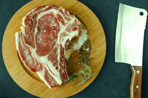 A slice of raw beef meat on the table with rosemary and other spices, rib eye steak ingredients with pepper and coarse salt