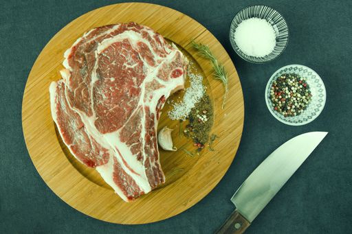 A slice of raw beef meat on the table with rosemary and other spices, rib eye steak ingredients with pepper and coarse salt