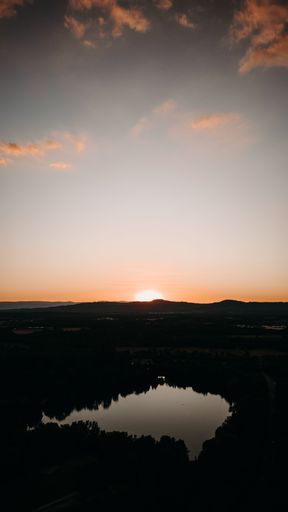 A vertical aerial view of a small lake in a park at sunset with an orange cloudy sky in the background