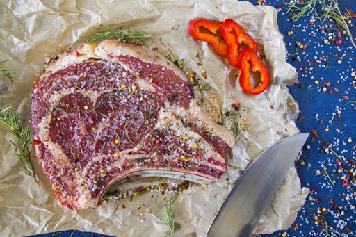 A slice of raw beef meat on the table with rosemary and other spices, rib eye steak ingredients with pepper and coarse salt