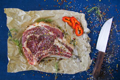 A slice of raw beef meat on the table with rosemary and other spices, rib eye steak ingredients with pepper and coarse salt