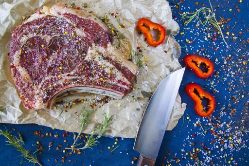 A slice of raw beef meat on the table with rosemary and other spices, rib eye steak ingredients with pepper and coarse salt