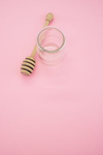 A top view closeup of a wooden honey dipper next to a glass container placed on a pink background