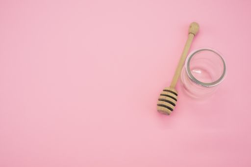 A top view closeup of a wooden honey dipper next to a glass container placed on a pink background