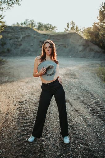A Bosnian Caucasian woman holding a cowboy hat while standing on an unpaved road in the countryside