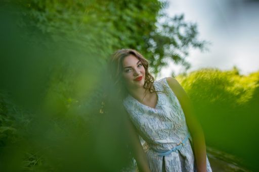 A portrait of a smiling sexy brunette girl in nature posing behind blur green leaves on a light summer day