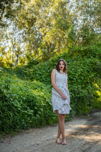A vertical shot of a brunette female holding her summer dress with a hand in the green plants' background