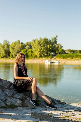 A full body shot of a young sexy brunette girl sitting on a rock by a small lake during sunrise