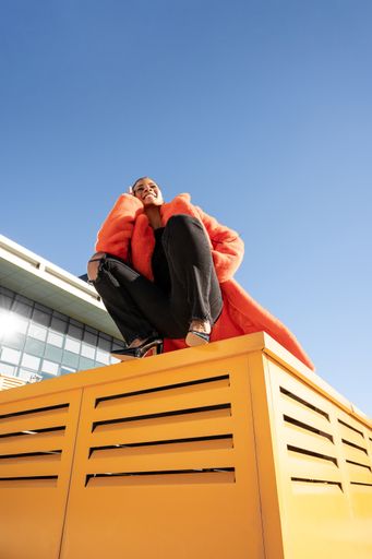 A happ black female with a red coat sitting on a yellow surface and smiling