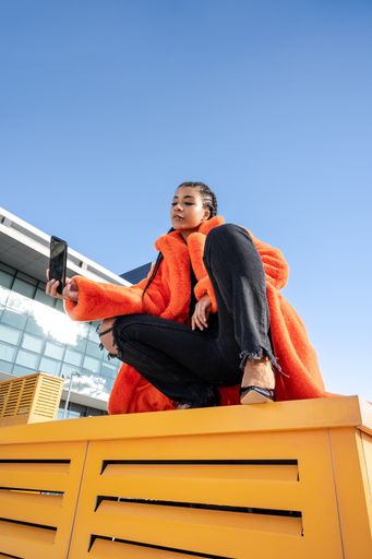 A young black female with a red coat sitting on a yellow surface and taking a selfie