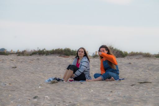 Two young pretty female friends sitting on a sandy beach pointing at something